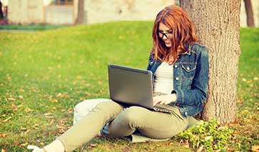 Student writing on a laptop leaning against a tree on a lawn