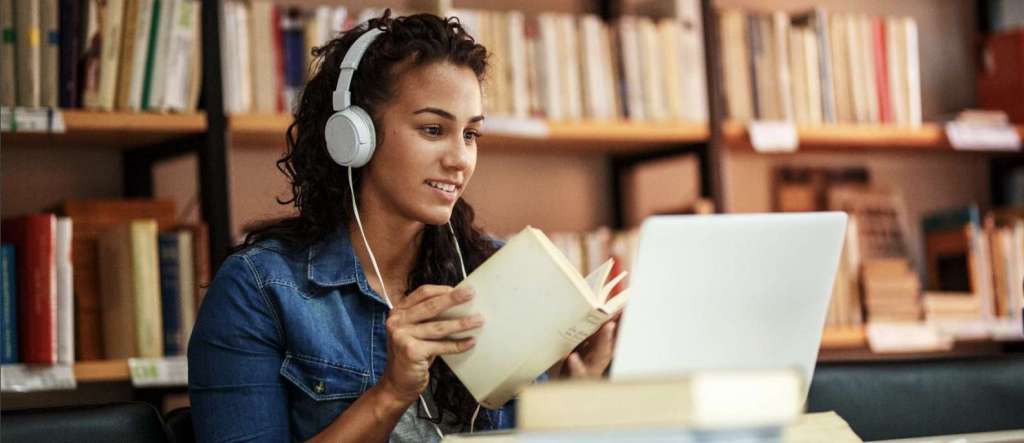girl with headphones on holding a book in front of a laptop